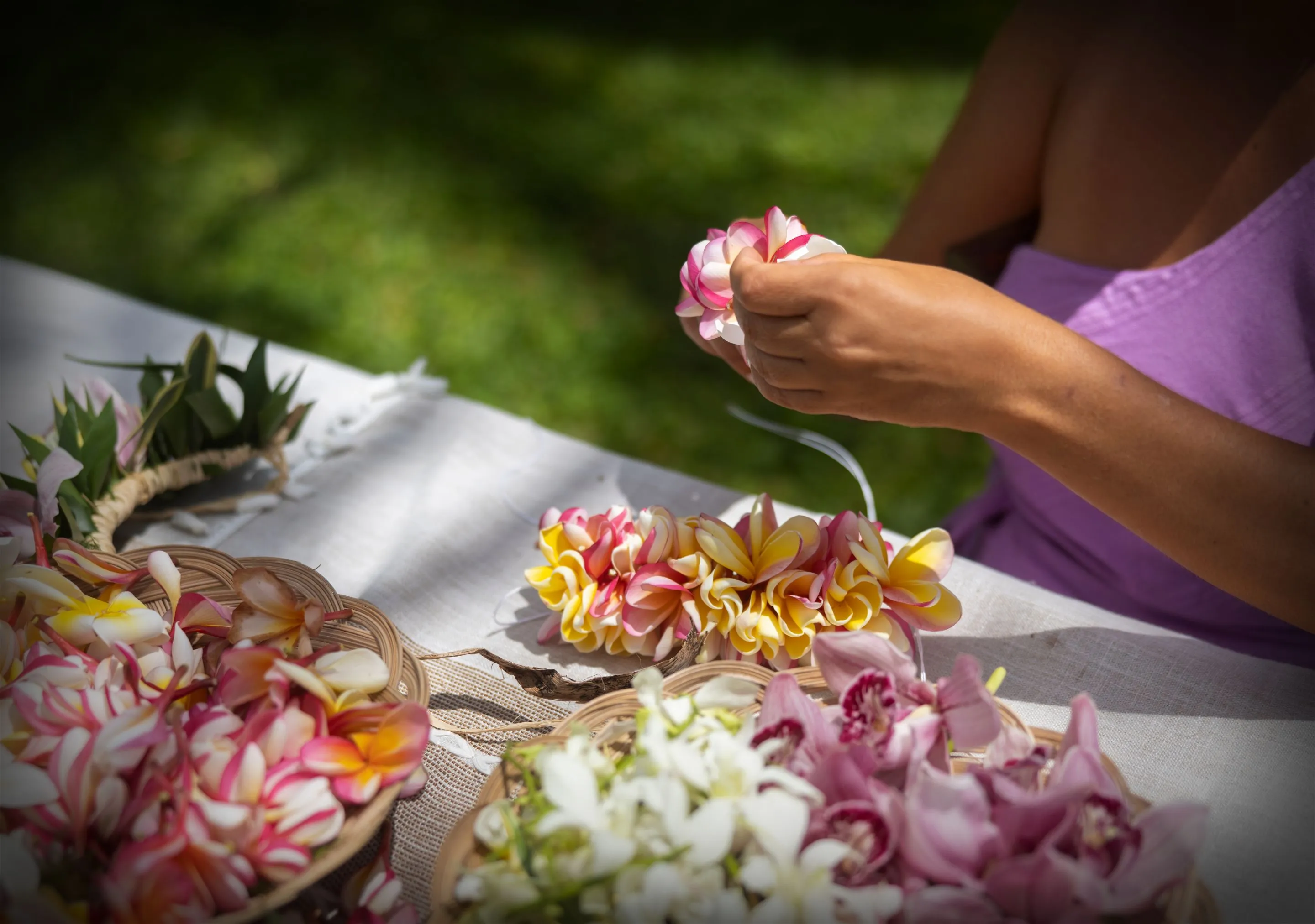 Person crafting floral leis at an outdoor table.
